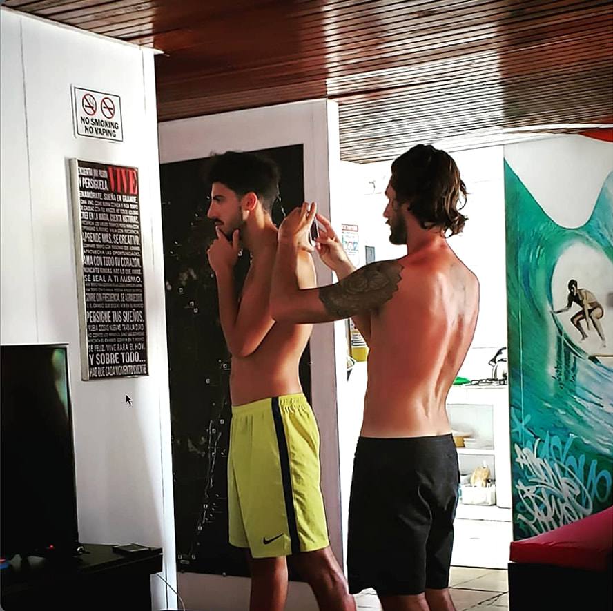 Two guests in the common room, one carefully photographing the other in front of the green surf mural.