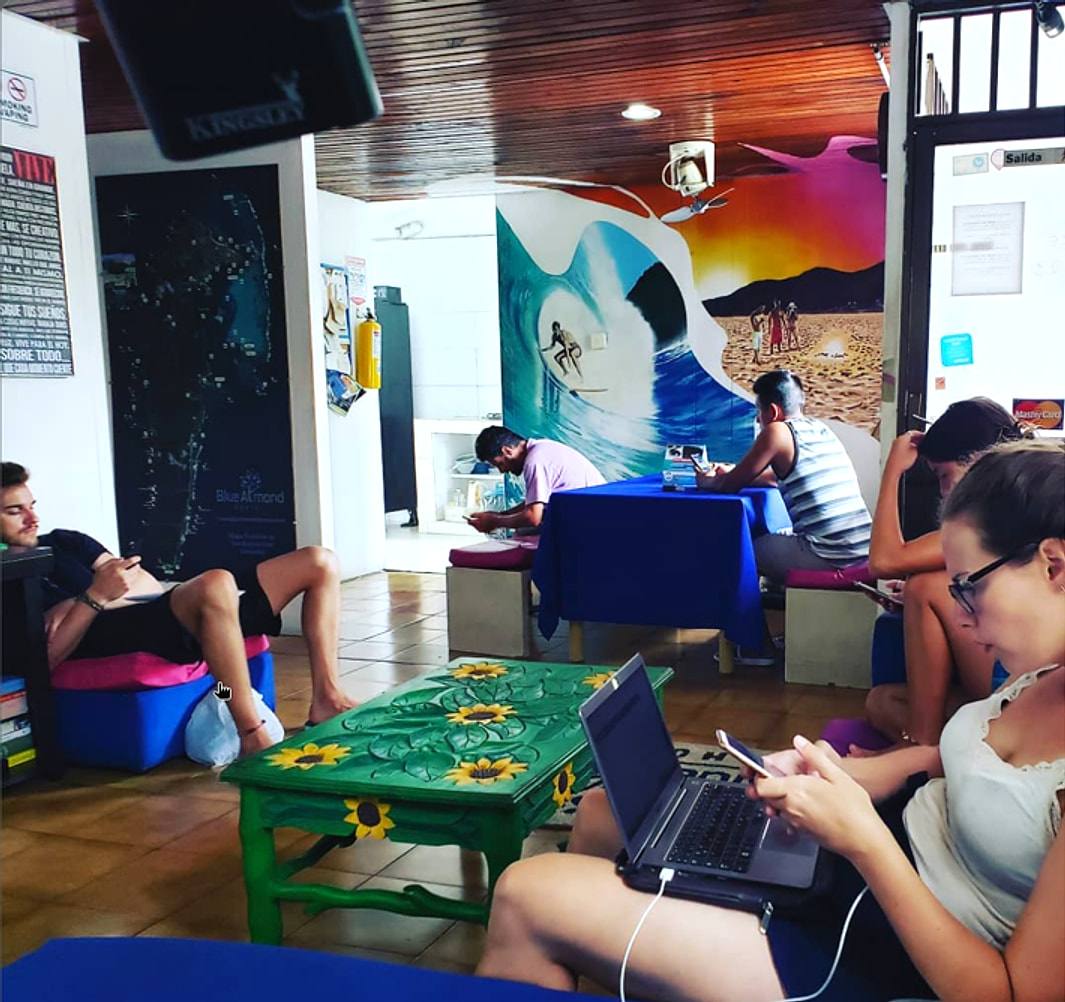 Blue Almond’s common room: guests on laptops and phones around a green table painted with sunflowers, under a huge turquoise wave mural with a surfer.
