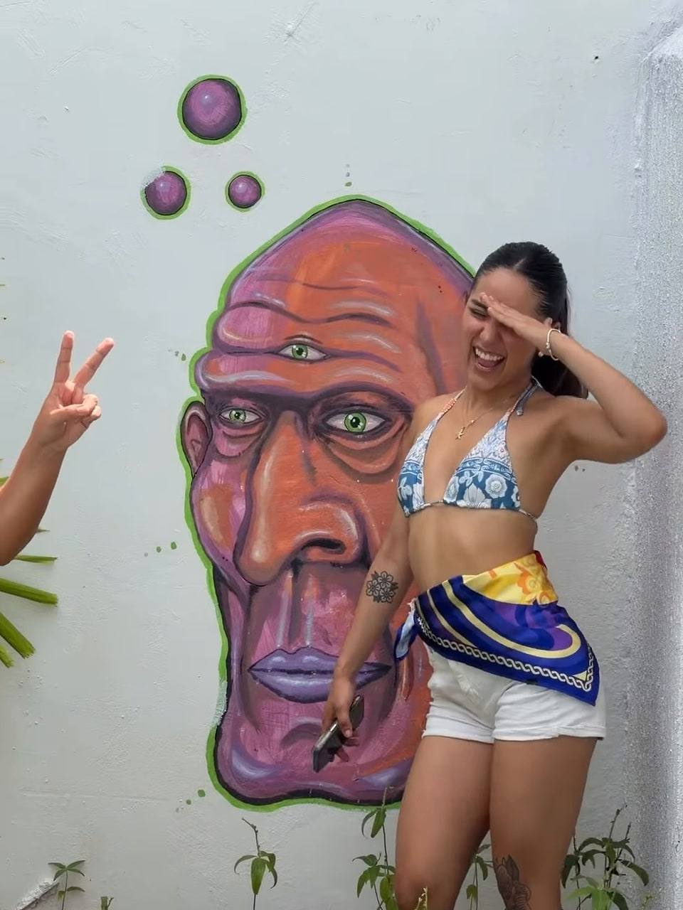 A guest posing next to a giant painted face with a third eye on the courtyard wall.
