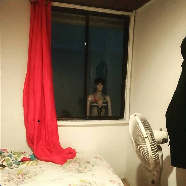 An old hostel room photo from 2019: bed, red curtain, standing fan and a mannequin nicknamed Stacy peering through the window.